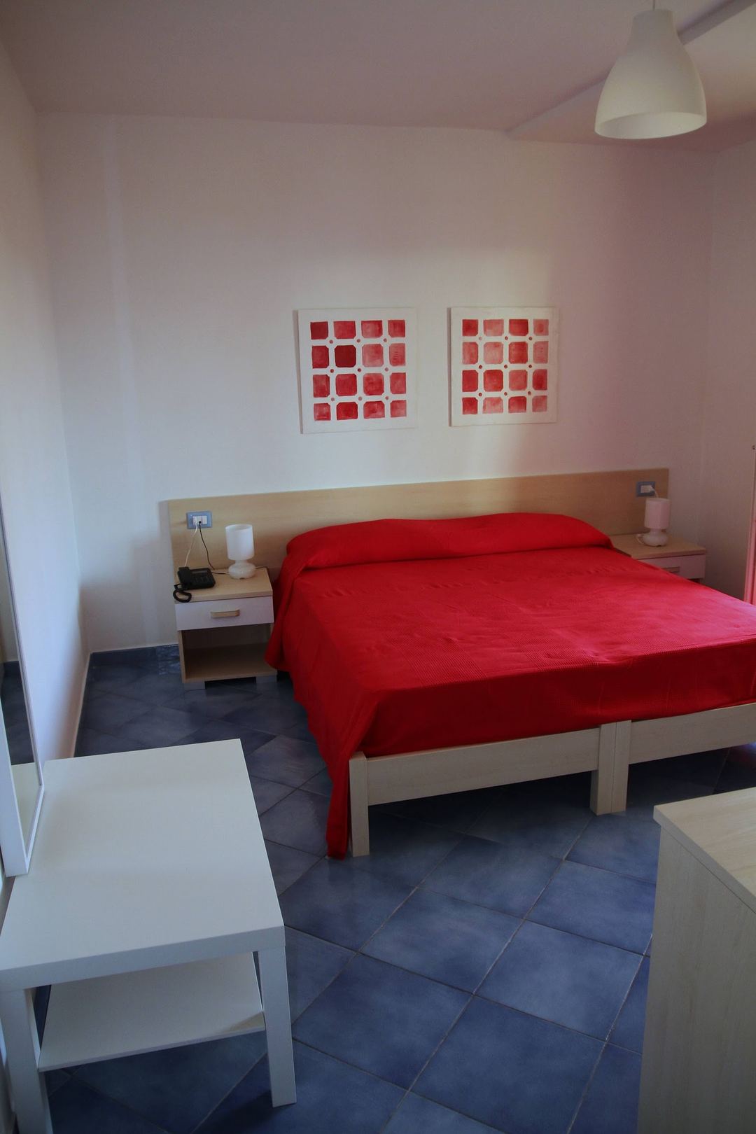 A modern bedroom featuring a bright red bed with wooden frame, white walls accented with pink, and minimalist furnishings including two red abstract artworks above the headboard.