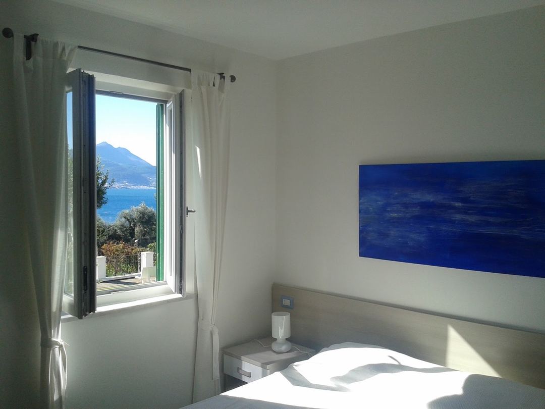 A bright bedroom with an open window overlooking a blue sea, mountain, and coastal landscape.
