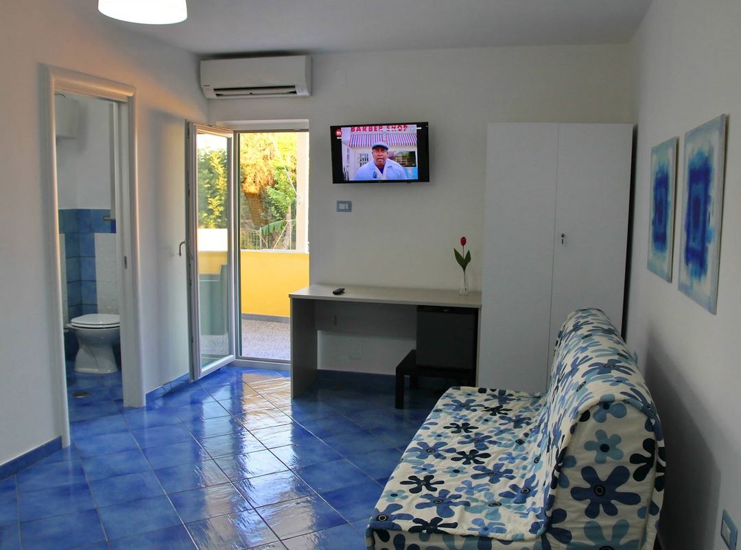 A bright guest room or common area featuring blue tile flooring, a desk, patterned sofa, and access to an outdoor terrace.