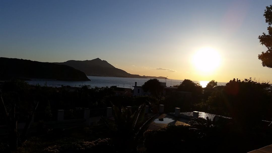 Sunset view over a coastal bay with silhouetted mountains and vegetation in the foreground.