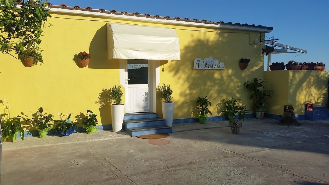 Bright yellow-painted entrance of B&B Belvedere with white awning, potted plants, and blue trim on a sunny day.