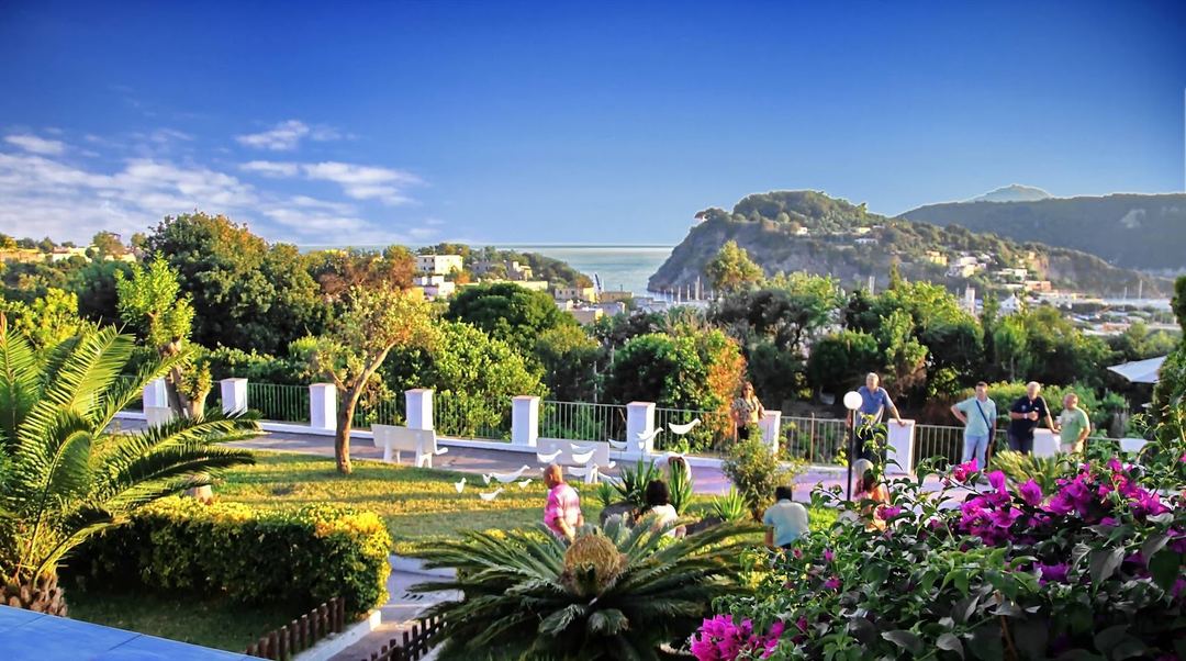 A scenic terrace garden overlooks a Mediterranean coastal town with mountains and a sparkling bay in the distance.