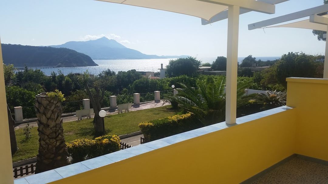 A scenic coastal view of a snow-capped mountain across the water, framed by a yellow-walled terrace with pergola and manicured gardens.